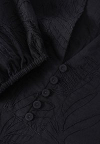 Tissu noir avec broderie à motif de feuilles et cinq boutons assortis le long du devant. Finition texturée avec des détails élastiques froncés.