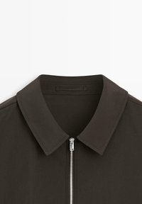 Donkerbruin shirt met een spitse kraag en een zilveren voorritssluiting, gefotografeerd vanaf de bovenkant van de borst tot aan de kraag tegen een witte achtergrond.