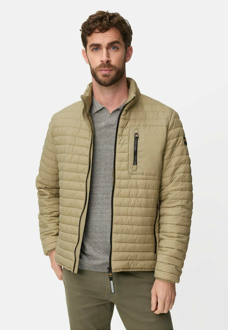 Mand med kort krøllet brunt hår iført en lys olivengrøn quiltet jakke over en grå skjorte og olivengrønne bukser, stående mod en hvid baggrund.