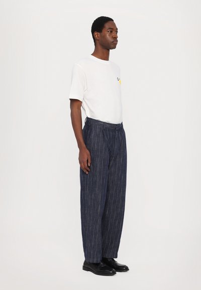 Witte t-shirt met korte mouwen en gele graphic, gecombineerd met donkerblauwe gestreepte broek en zwarte schoenen, gedemonstreerd door een model.