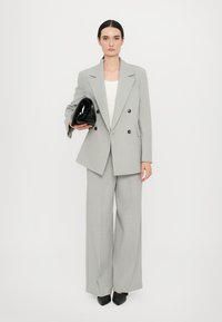 Lys grå, dobbeltradet dressjakke med svarte knapper, sammen med vide bukser. Holder en svart clutch, iført spisse svarte hæler.
