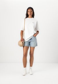 White oversized T-shirt with a small logo, paired with light blue denim shorts. Accessories include a beige shoulder bag and white sneakers.