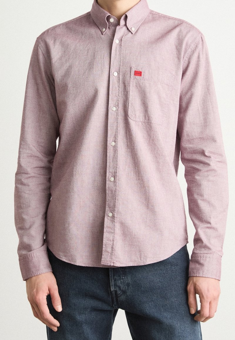 Homme portant une chemise violette claire à boutons avec une poche poitrine et un jean, debout avec les bras détendus.