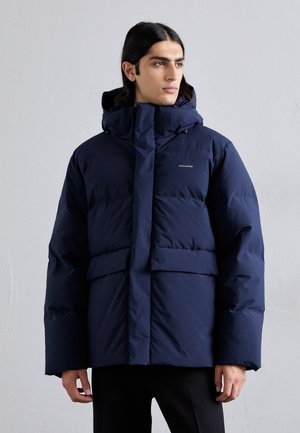 Chaqueta acolchada de color azul marino con un cuello alto, capucha, dos bolsillos frontales y un logo de marca. Presenta un corte relajado y una textura suave.