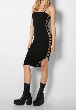 Svart ribbad klänning med tunna axelremmar, asymmetrisk nederdel och utklippta sidodetaljer. Har silverdetaljer längs sidan, i kombination med svarta plattformstövlar.