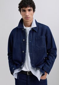 Jonge man in een donkerblauwe denimjack, lichtgestreept shirt, witte T-shirt en zwarte riem, met zijn handen in zijn zakken, kijkend naar voren.