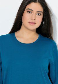 Tee mit langen Ärmeln in Tealfarbe, Rundhalsausschnitt, weicher Textur und subtiler Rippstruktur. Hat einen lässigen Schnitt und keine sichtbaren Beschläge.