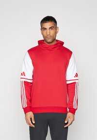 Röd sweatshirt med högt halsliv, med vita ärmar och tre vita ränder på varje arm. Materialet verkar mjukt och strukturerat.