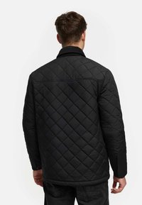Chaqueta negra acolchada con patrón de diamantes, con cuello puntiagudo y mangas largas. El material texturizado proporciona aislamiento.