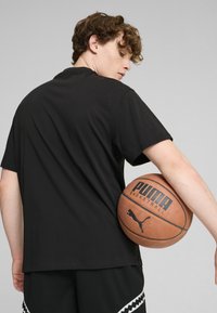 Čierne bavlnené tričko s krátkymi rukávmi, nosené s rubom von, v kombinácii s čiernymi sieťovanými šortkami. Basketbalová lopta s brandingom Puma je opretá o ruku.