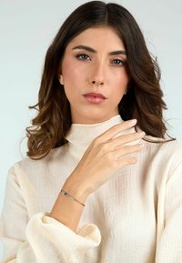 Blusa di colore crema con texture e colletto alto, indossata con un bracciale argento dal design minimale. Capelli ondulati e di lunghezza alla spalla.