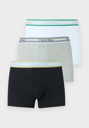 Hemdeloze man die blootsvoets staat en lichtgrijze boxershorts draagt met een Calvin Klein-liesband op een effen witte achtergrond.