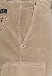 Beige kortkottsbyxor med en texturerad yta, som har en framficka, svarta metalldetaljer och en liten varumärkesetikett.