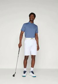 Moška golfskaoutfit, ki vključuje modro polo majico, bele hlače, svetlo pas in bele superge z modrimi detajli, ki drži golf palico.