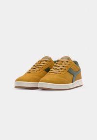 Baskets en daim jaune avec des accents verts, semelle en caoutchouc blanche et lacets beige. Caractéristiques d'une texture lisse et de détails de couture contrastants.