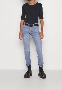 Kvinna i svart knäppt topp, ljusblå slitna jeans med svart spikad bälte, och svarta plattformsskor med snörning.