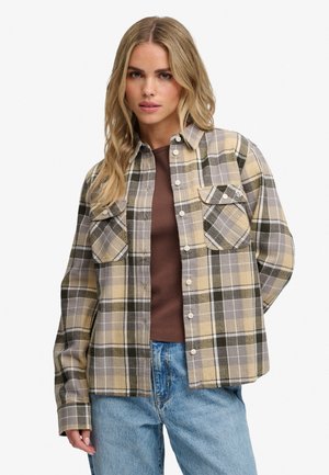 Ung kvinde med langt blondt hår iført en beige og grå ternet skjorte over en brun top og lyseblå jeans.