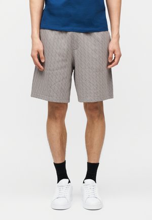 Man wearing gray patterned shorts, black socks, white sneakers, and a blue shirt standing against a plain light background.