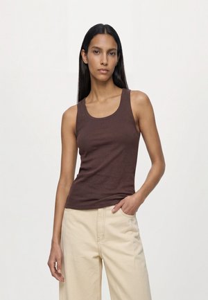Femme aux longs cheveux noirs portant un haut marron sans manches et un pantalon beige clair, debout avec une main dans la poche sur un fond uni.