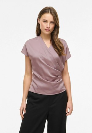 Femme portant une blouse en satin mauve style portefeuille et un pantalon large noir, debout devant un fond blanc uni.