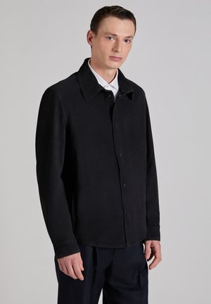 Młody mężczyzna w czarnej, zapiętej na guziki kurtce nałożonej na białą koszulę, stoi na jednolitym jasnoszarym tle.