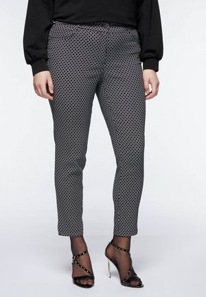 Nainen, jolla on mustavalkoiset kuvioidut kapeat housut, mustat läpikuultavat sukkahousut, mustat remmikorolliset sandaalit ja musta pitkähihainen paita.