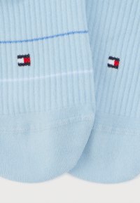 Φωτεινά γαλάζιες ριγωτές κάλτσες με μικρά κόκκινα, λευκά και μαύρα ορθογώνια λογότυπα κοντά στα δάχτυλα, τοποθετημένες δίπλα-δίπλα σε λευκό φόντο.