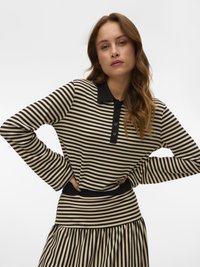 Donna con capelli lunghi che indossa una maglietta a righe orizzontali beige e nera con colletto e bottoni neri, mani sui fianchi, sfondo neutro.