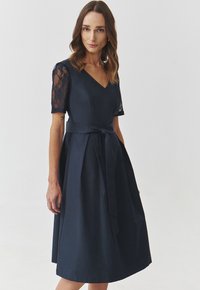 Navyblaues Kleid mit V-Ausschnitt, kurzen Spitzenärmeln und tailliertem Oberteil. Hervorhebung einer gebundenen Taille und eines fließenden Rocks, glatte Stofftextur.
