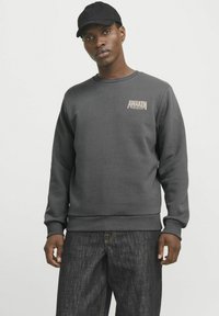 Grå sweatshirt med ribbede manchetter, der har et lille "AWAKEN" tekstlogo. Sammen med mørke denimjeans og en sort kasket.