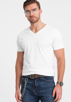 Vit V-ringad t-shirt i bomull, med korta ärmar. Bärs med blå jeans och ett mönstrat bälte. Accessoarer inkluderar en klocka och glasögon.