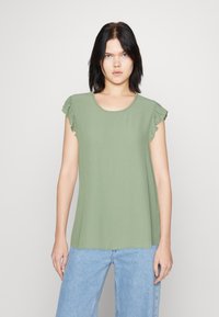 Haut vert à manches courtes avec des accents volantés, col rond et tissu doux ; associé à un jean large bleu clair.