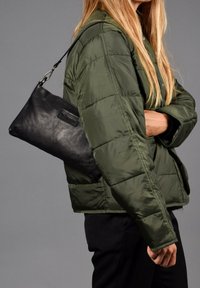 Bolso de mano negro de piel con un acabado texturizado, que cuenta con una cremallera superior y una correa desmontable, que se lleva debajo de una chaqueta de abrigo verde.