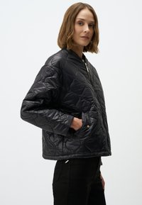 Veste noire matelassée avec fermeture éclair sur le devant, col arrondi et poches latérales avec des accents en métal doré. Matériau lisse et brillant.