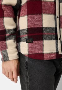 Rød og hvid ternet fleece-skjorte med sorte detaljer, knappelukning og sort mærke med teksten "made for adventures" på den nederste kant.