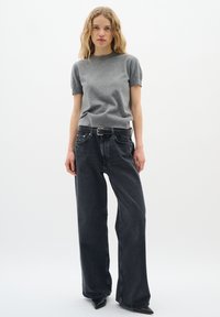 Frau mit welligem blonden Haar, die einen grauen Kurzarmpullover, schwarze weit geschnittene Jeans mit einem schwarzen Gürtel und spitze schwarze Schuhe vor einem weißen Hintergrund trägt.