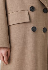 Cappotto beige su misura con tessuto texturizzato, design doppiopetto e bottoni neri. Include un dettaglio con tasca piatta sul lato.