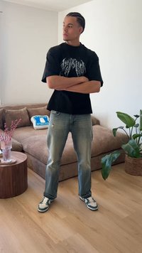 Homme aux cheveux tressés, debout les bras croisés, portant un t-shirt noir à motif, un jean clair et des baskets dans un salon avec des plantes et un canapé.