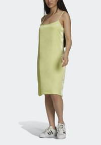 Kobieta w żółtej sukience na ramiączkach i białych sneakersach, stojąca na tle jednolitego jasnego tła.