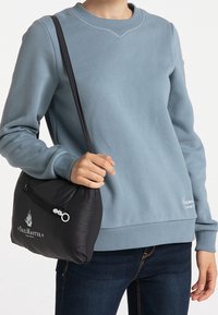 Een zwarte, zachte handtas met een rits bovenkant, met een logo, vastgehouden door een persoon in een lichtblauwe sweater en donkere jeans.