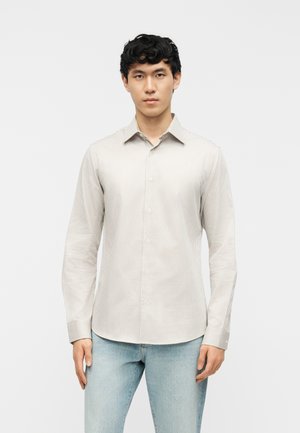 Mladý muž s krátkymi kučeravými vlasmi, oblečený v béžovo-bielej vertikálne pruhovanej košeli na gombíky a svetlomodrých džínsoch, stojaci pred jednoliatym pozadím.