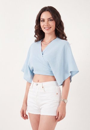 Giovane donna con capelli castani mossi che indossa un top corto azzurro chiaro e pantaloncini bianchi, sorridente, in piedi davanti a uno sfondo bianco uniforme.
