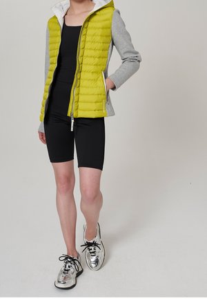 Person iført en gul polstret vest med grå ærmer, sort ærmeløs top, sorte cykelshorts og sølvfarvede sneakers med sorte snørebånd.