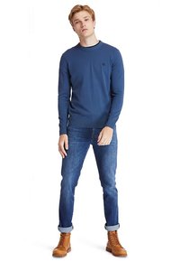 Niebieski sweter z dekoltem typu crewneck z prążkowanym wykończeniem, zestawiony z podwiniętymi jeansami i brązowymi sznurowanymi butami. Prosty detal z logo na swetrze.