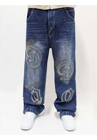 Jeans de mezclilla azul con ajuste suelto, que presentan intrincados patrones de bordado de color claro y una textura desgastada. Se llevan zapatillas blancas.