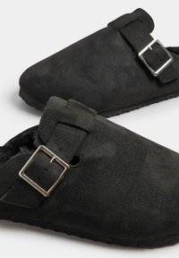 Zuecos de ante negro con una sola correa de hebilla, suela plana y un acabado suave y texturizado. Diseñados para ofrecer comodidad y facilidad de uso.