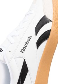 Zapatilla blanca de Reebok con detalles en cuero negro, suela de goma texturizada y cordones blancos. Presenta costuras decorativas y un logo.