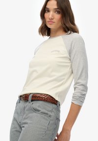 Vrouw draagt een beige en grijs shirt met lange mouwen en een "Superdry" logo, gestreepte broek en een bruine gevlochten riem, staand tegen een witte achtergrond.