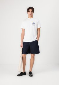 Hvit bomullst-skjorte med et blått grafisk design og tekst, kombinert med mørke denimshorts. Modellen holder en beige totebag og har på seg svarte sko.