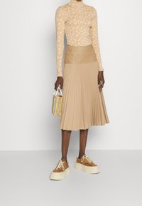 Beige långärmad topp med blommönster, beige plisserad kjol och bruna platåskor. Håller en liten rutig handväska.